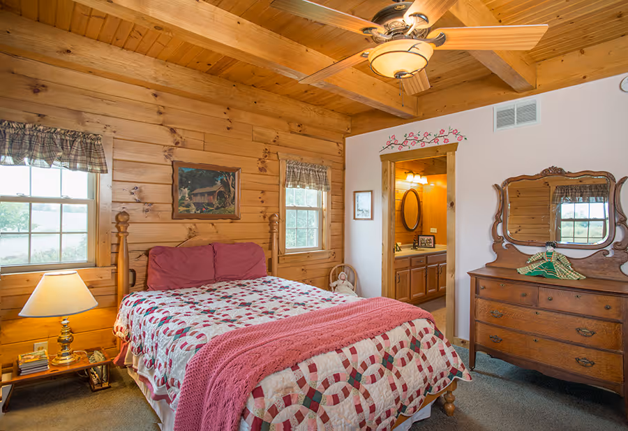A cozy wooden bedroom featuring a double bed with a quilted red and white coverlet, two windows with green curtains, and a wooden dresser beside a lamp. Decor includes a wall painting and an open door leading to a bathroom.