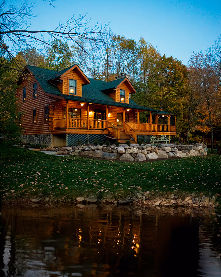 A charming wooden cabin with a green roof situated by a serene lake, surrounded by autumn trees. The cabin features multiple balconies and warm lights reflecting off the water in the evening glow.