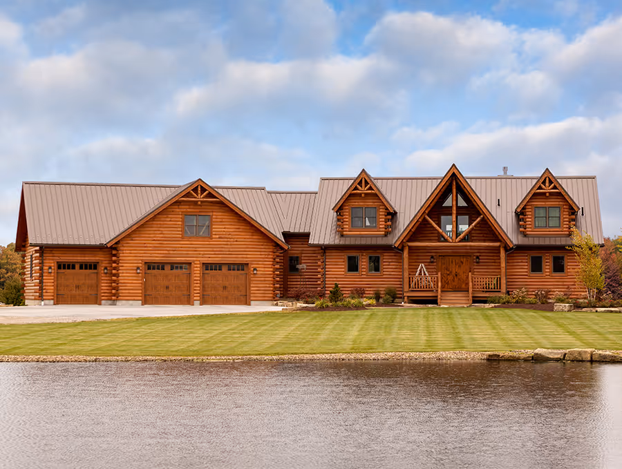 A spacious log cabin home with a large front lawn, two garage doors, and a distinctive triangular window design. The home is set against a serene pond and a backdrop of soft, cloudy skies.