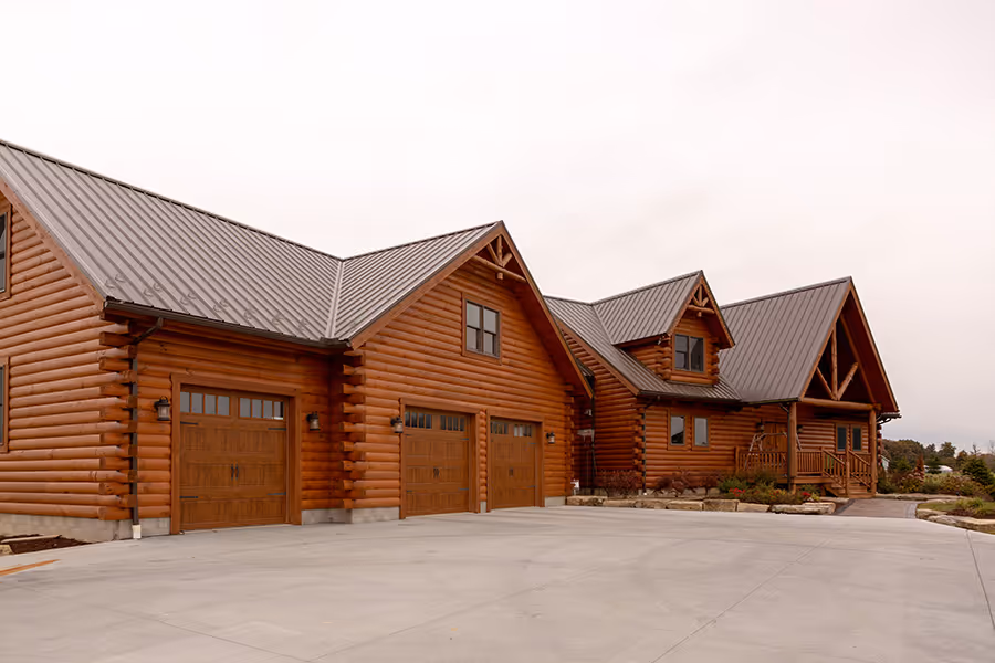 A spacious log cabin with wood exterior and a sloped metal roof, featuring multiple gabled sections and large garage doors. The driveway is paved and leads to a landscaped area in front of the house.