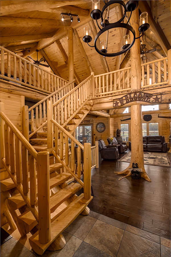 Interior view of a cozy log cabin featuring wooden spiral staircases and a rustic chandelier. The warm wood tones create an inviting atmosphere, complemented by comfortable seating in the living area.