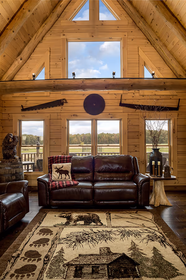 A cozy cabin living room with wooden walls and a vaulted ceiling, featuring a brown leather sofa adorned with a plaid pillow. A large area rug with nature motifs covers the floor, and panoramic windows provide a view of greenery outside.