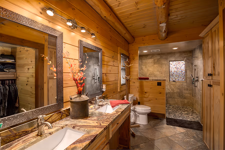 A cozy bathroom featuring wooden walls and ceiling, illuminated by ceiling lights. The vanity has a marble sink and a decorative vase with flowers. A large mirror reflects a contemporary shower area with stone tiles and a colorful glass panel.