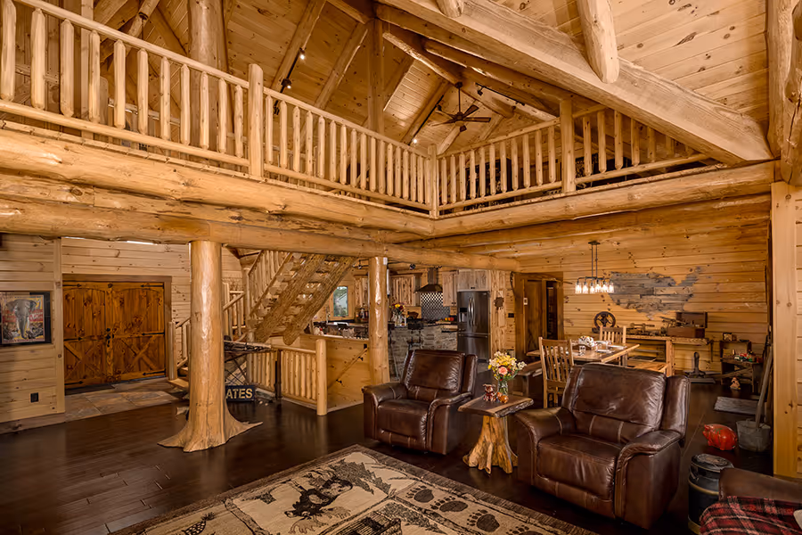 A spacious log cabin interior featuring exposed wooden beams and a cozy seating area with brown recliners. A staircase leads to a loft area, while a dining space and kitchen are visible in the back. A colorful area rug adds warmth to the wooden floor.