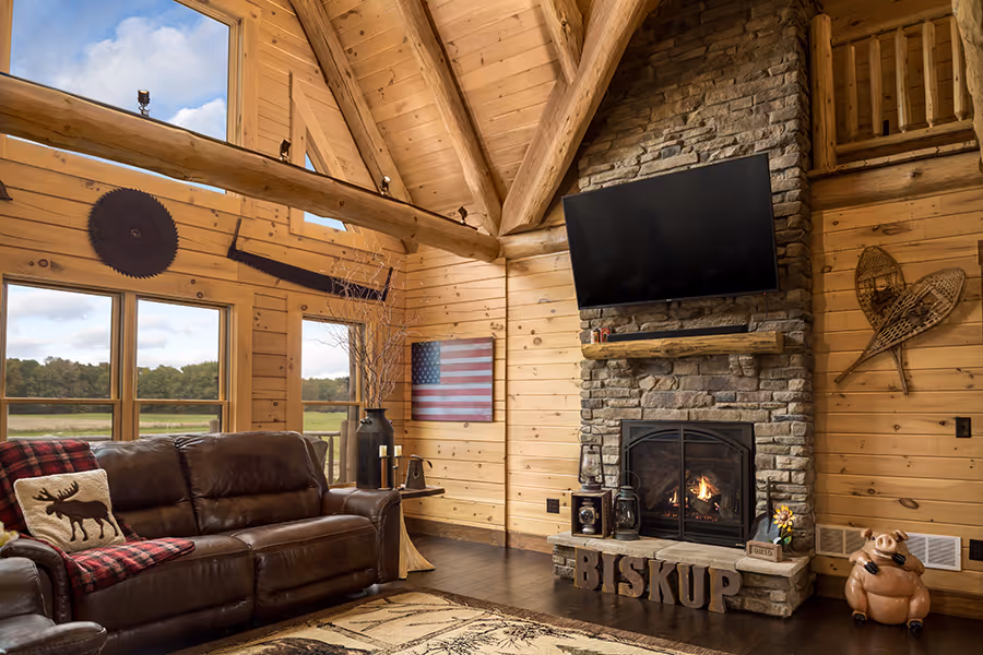 A rustic living room featuring a large stone fireplace, a leather couch with a red plaid throw, and large windows overlooking a green landscape. The wooden walls and beams add warmth, while a TV is mounted above the fireplace.