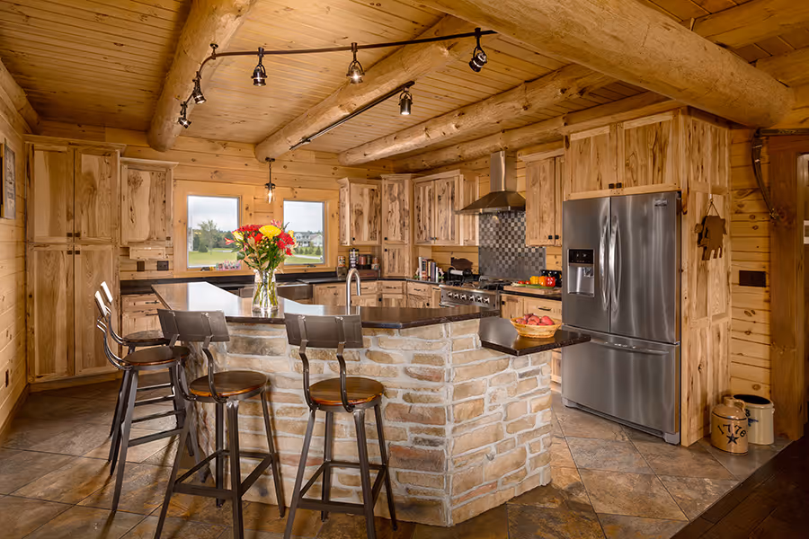 A rustic kitchen featuring wooden cabinets, a stone island, and modern appliances. A vase with red flowers decorates the island, while bar stools provide seating. Large windows allow natural light to illuminate the space.
