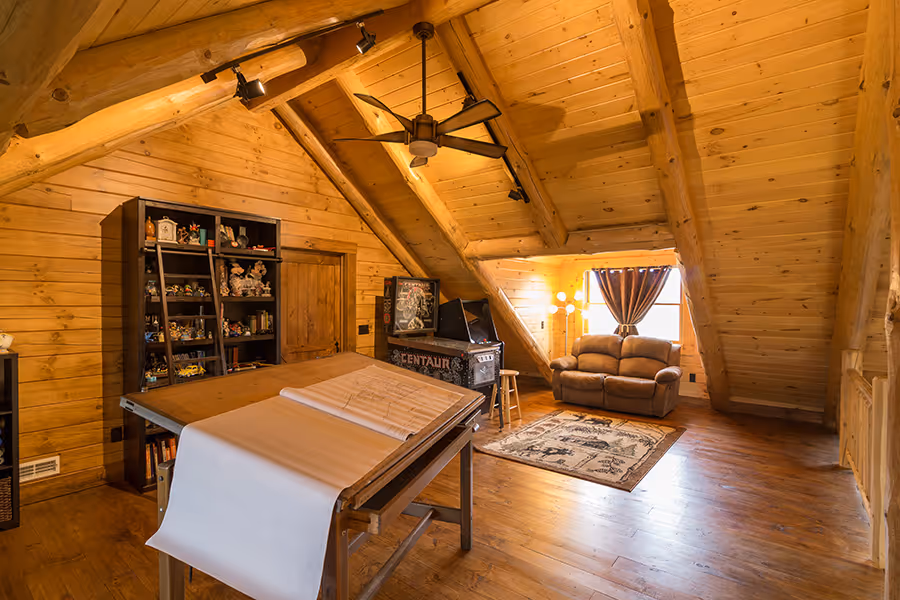 A cozy attic office with wooden beams and warm lighting. A large, light wooden table is positioned in the center, holding rolled-up drawing paper. A comfortable seating area with a brown couch and a side table is near a window, adorned with soft curtains.