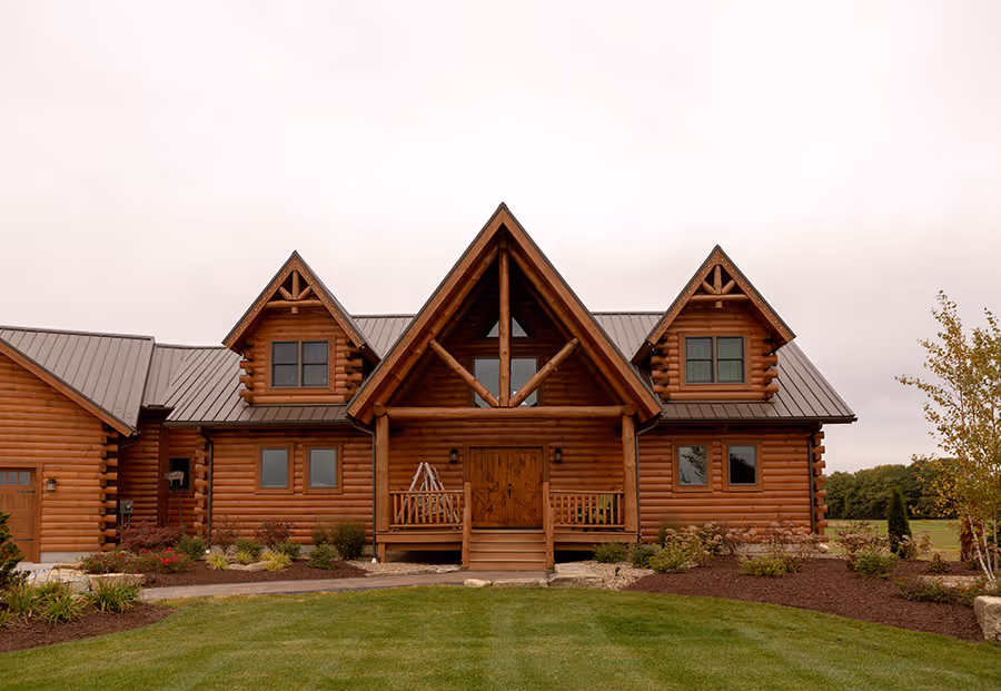 A large, rustic log cabin with a steep triangular roof, featuring large windows and a welcoming front porch. The home is surrounded by a well-kept lawn and landscaped with shrubs and small trees, showcasing a serene outdoor setting.