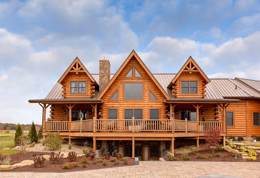 A large log cabin house featuring multiple gabled roofs, a wooden deck, and large windows, surrounded by landscaped gardens and a paved driveway, under a cloudy sky.