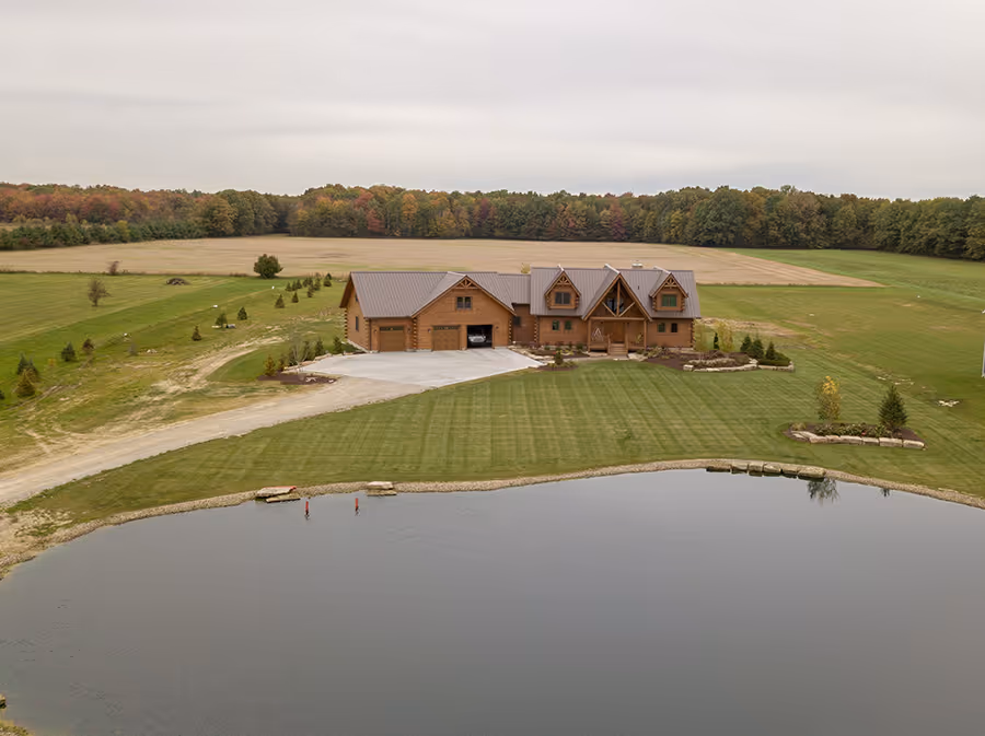 Aerial view of a large wooden house with a sloped roof, surrounded by lush green lawns and a calm pond reflecting the sky. Trees in the distance showcase fall colors with shades of green, orange, and yellow.