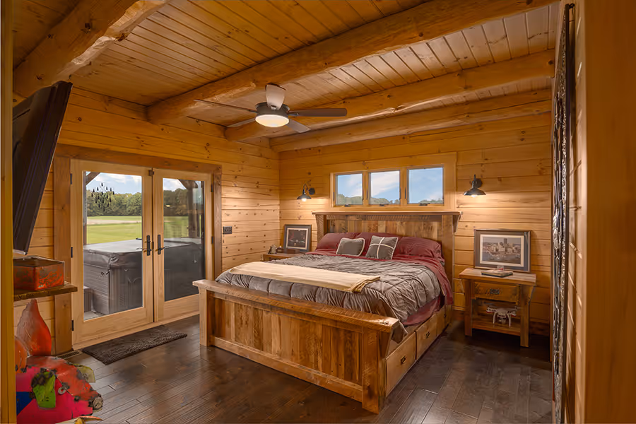 A cozy wooden bedroom featuring a large bed with a thick comforter, flanked by bedside tables. Warm wooden walls and floors create a rustic ambiance, with large glass doors opening to a scenic view outside. A ceiling fan adds comfort.