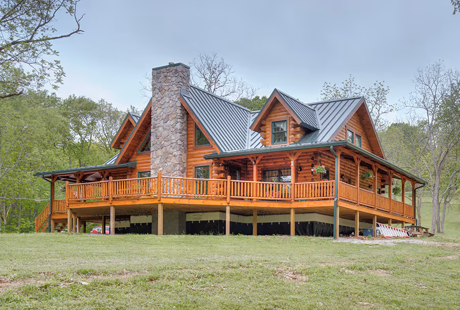 A large, rustic log cabin with a stone chimney and a sloped metal roof, featuring a spacious wraparound porch and surrounded by lush greenery. The building showcases a blend of wood and stone materials, giving it an inviting, natural appearance.