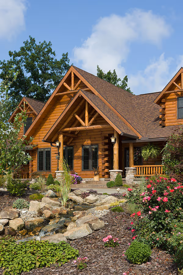 A charming log cabin home featuring a steep sloped roof and decorative wooden beams. The pathway leads to a warmly lit entrance, surrounded by vibrant flower beds and lush greenery. A small water feature adds a serene touch to the landscape.