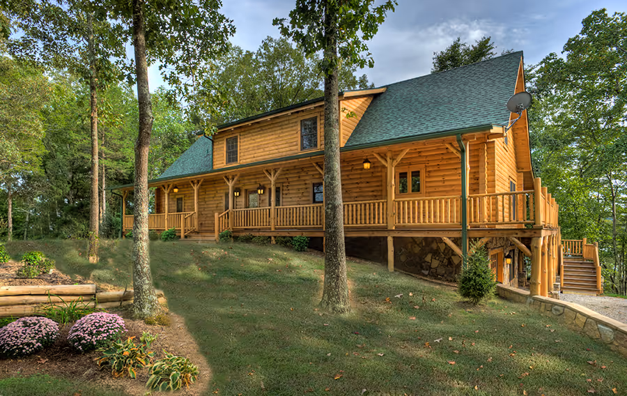 A rustic wooden cabin with a sloped green roof, surrounded by trees. The spacious porch features wooden railings, and colorful flowers bloom in the foreground. The cabin is set in a natural, green environment, showcasing a tranquil home backdrop.