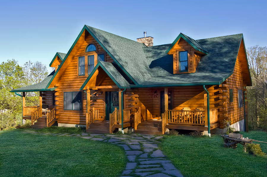 A beautiful log cabin with green roof and wooden porches, surrounded by a lush lawn and trees. The structure features large windows, a stone chimney, and a winding stone pathway leading to the front door.