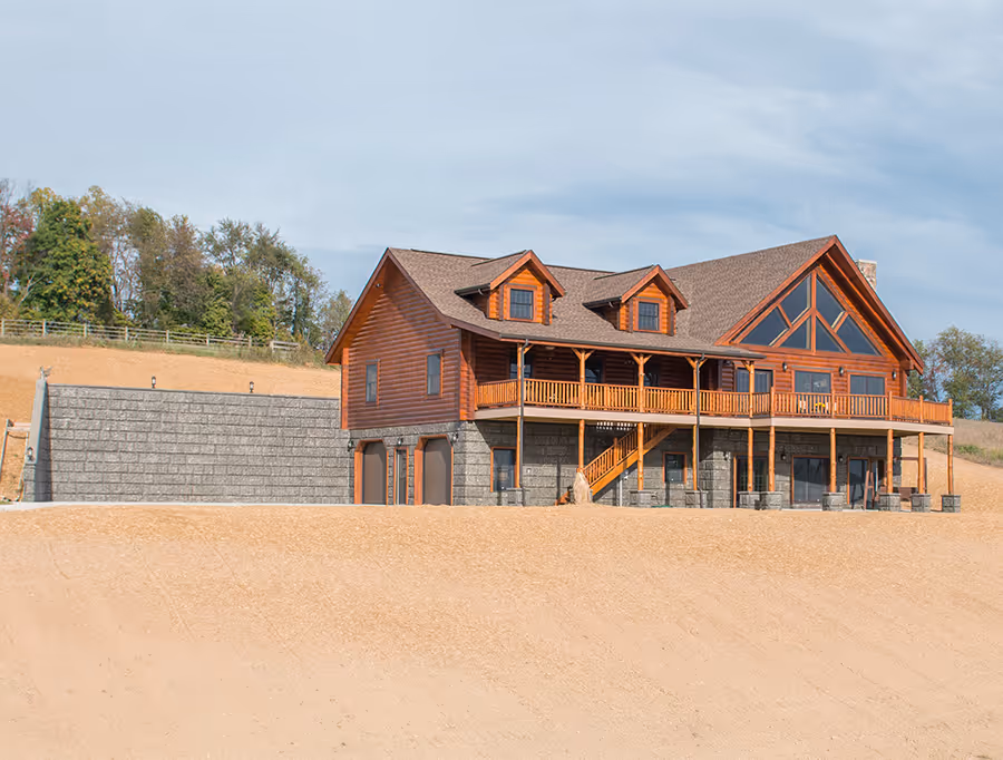 A large wooden lodge with a spacious deck and gabled roof, surrounded by natural landscape. The lodge features stone accents and large windows, offering a rustic yet modern architectural style. A sandy area is visible in the foreground.