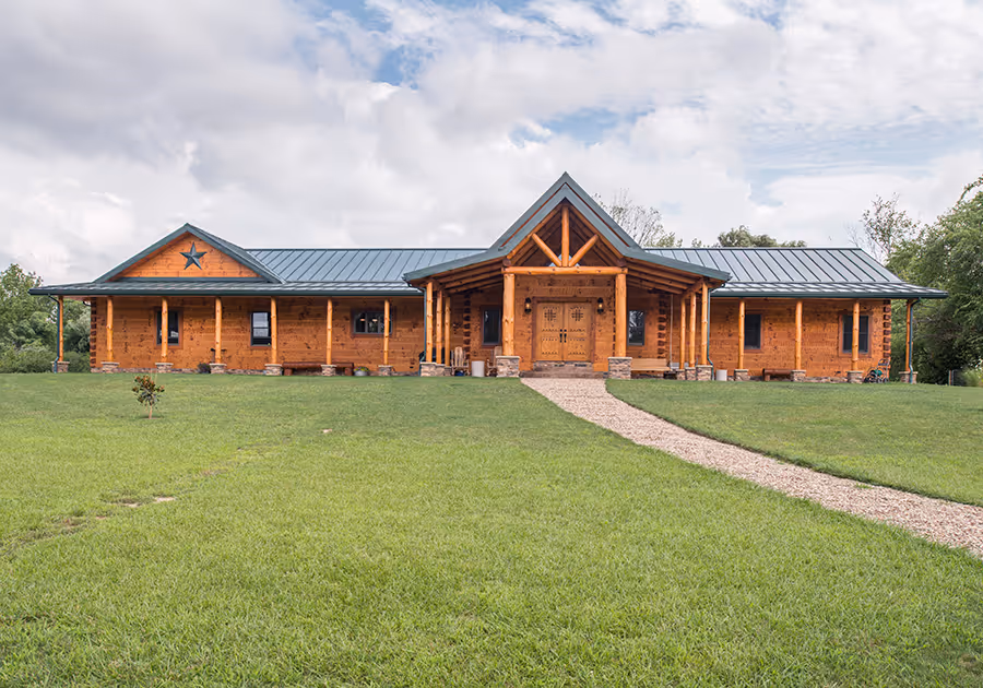 A large, wooden ranch-style house with a metal roof, featuring a star motif above the entrance and surrounded by lush green grass. A gravel path leads to the front door, with trees in the background under a cloudy sky.
