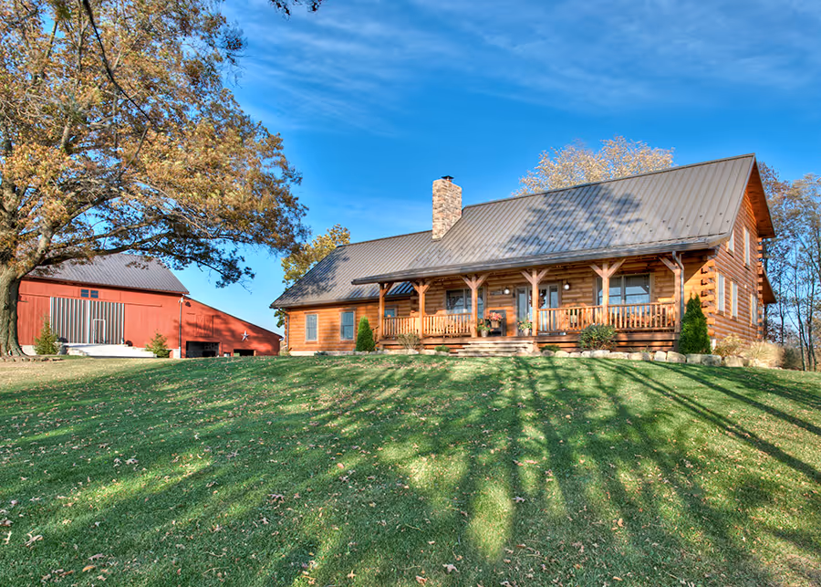 A charming log cabin with a covered porch and a gray metal roof, surrounded by a lush green lawn and tall trees. A red barn is visible in the background under a clear blue sky, enhancing the rustic countryside ambiance.