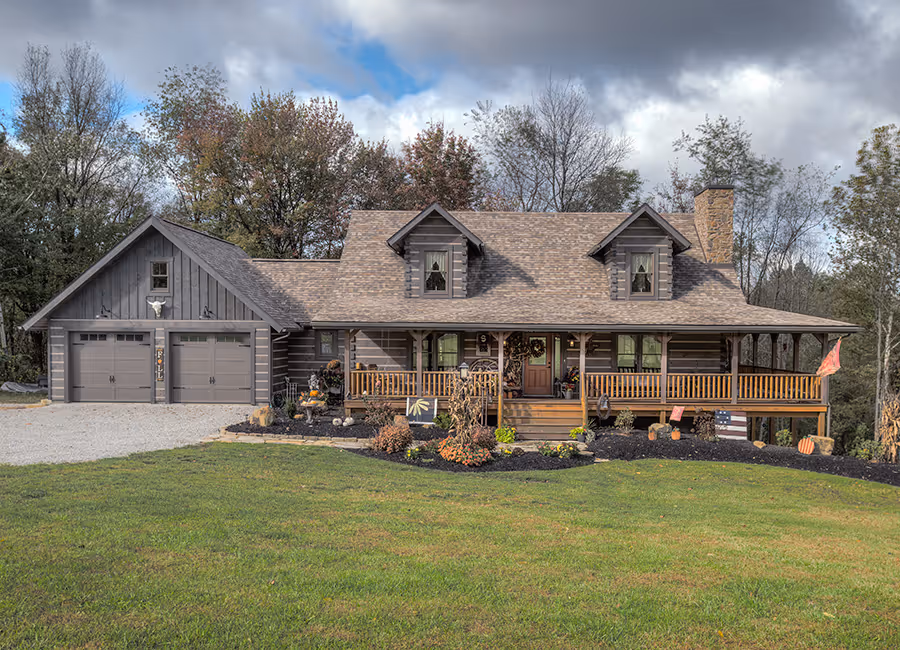 A charming log cabin-style house with a steeply pitched roof and a spacious front porch is surrounded by a lush green lawn and trees. The property features a gravel driveway and colorful flowerbeds, adding warmth to the picturesque setting.