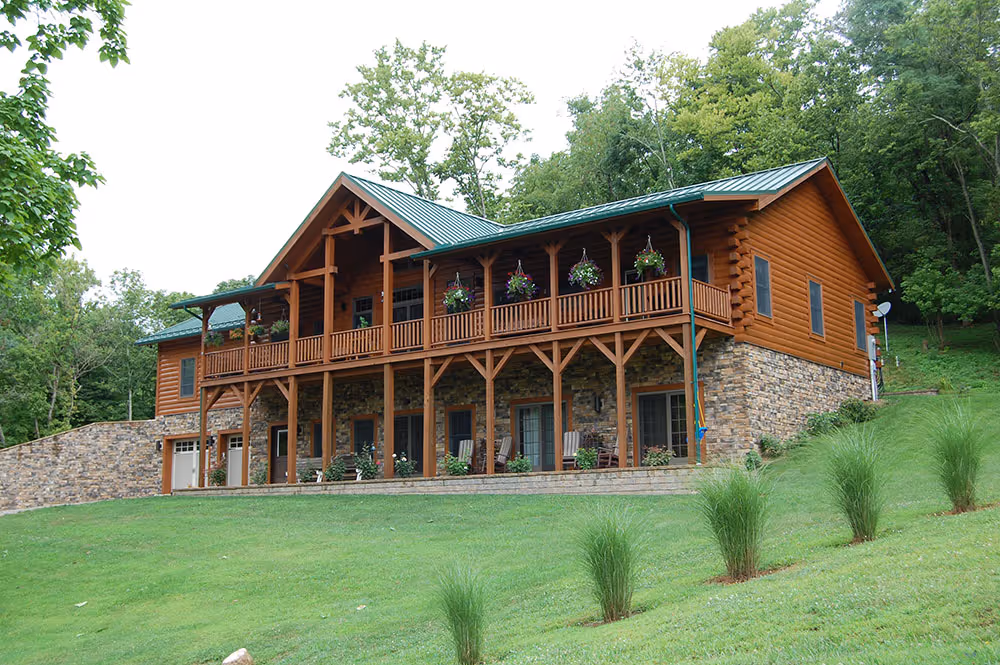 A large rustic cabin made of logs and stone, featuring a green metal roof and large porches adorned with hanging flower baskets. The house is surrounded by lush greenery and is set on a gently sloping hill.