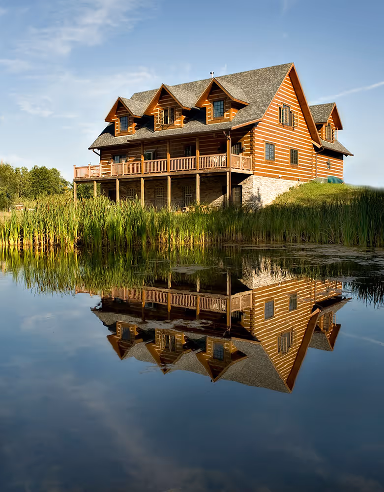 A large wooden lodge with multiple gabled roofs, surrounded by lush greenery and tall reeds, reflected in a calm lake under a clear blue sky.