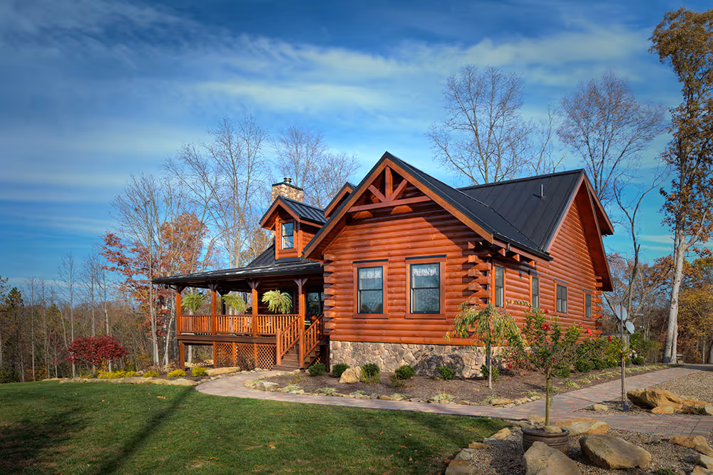 A charming log cabin featuring a dark roof and wooden siding, positioned amidst trees and greenery. The porch extends along the front, adorned with potted plants. A winding stone pathway leads to the entrance, under a clear blue sky.