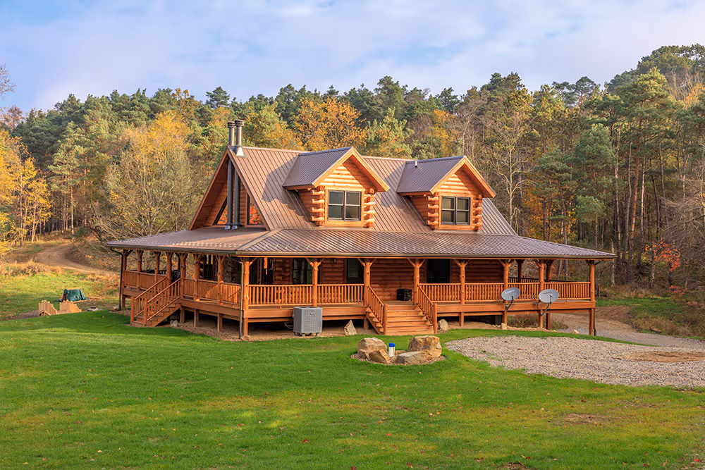 A spacious log cabin with a wide porch, surrounded by green grass and tall trees in the background. The cabin features a steep roof and multiple windows, providing a cozy and inviting atmosphere. The scene is set in a natural, wooded area during autumn.
