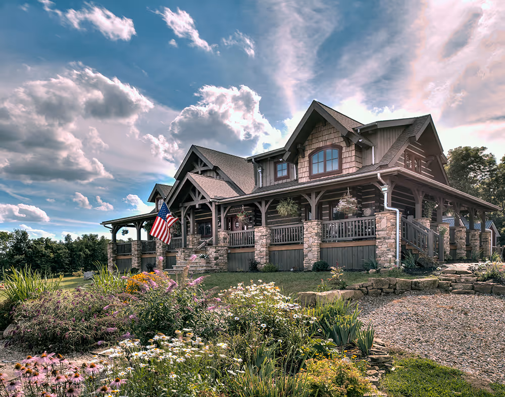 A beautiful log cabin-style house with a large porch, surrounded by colorful flowers and greenery, under a bright blue sky with fluffy white clouds. An American flag is proudly displayed on the porch.