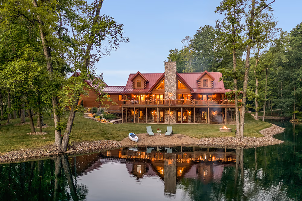 A large wooden cabin with a red metal roof situated by a serene pond, surrounded by green trees. The cabin features a stone chimney, large windows, and a deck with chairs overlooking the water.