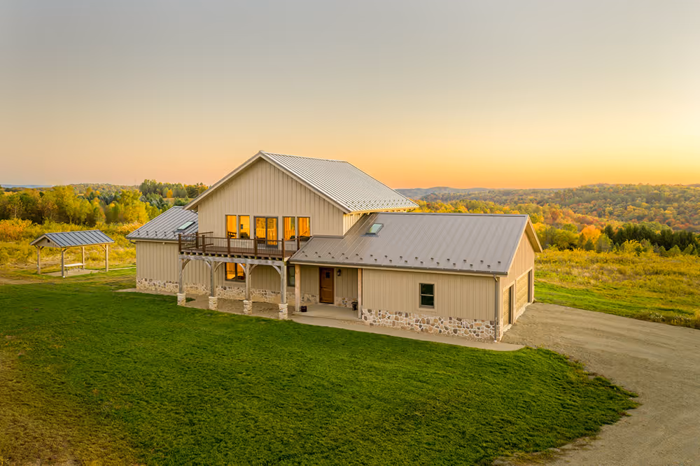 A modern rustic house with a light-colored exterior, large windows reflecting a sunset, and a surrounding green lawn. Trees and rolling hills are visible in the background, creating a serene landscape.