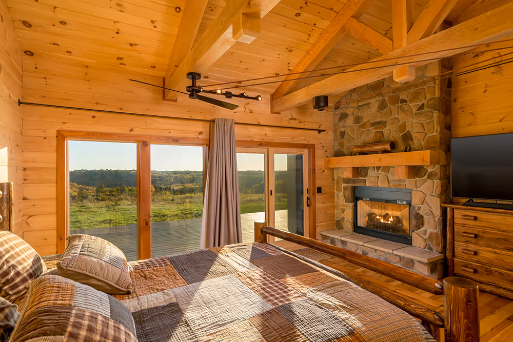 A cozy bedroom featuring a wooden bed with a plaid blanket, positioned beside a stone fireplace. Large windows allow natural light and showcase a scenic view of green hills outside. Warm wood paneling adds a rustic charm.