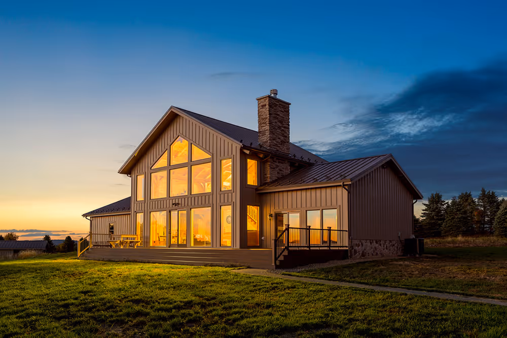 A modern house with large windows illuminated by sunset, showcasing a beautiful facade. The home is surrounded by lush green grass and a clear sky in the background, enhancing its tranquil setting.