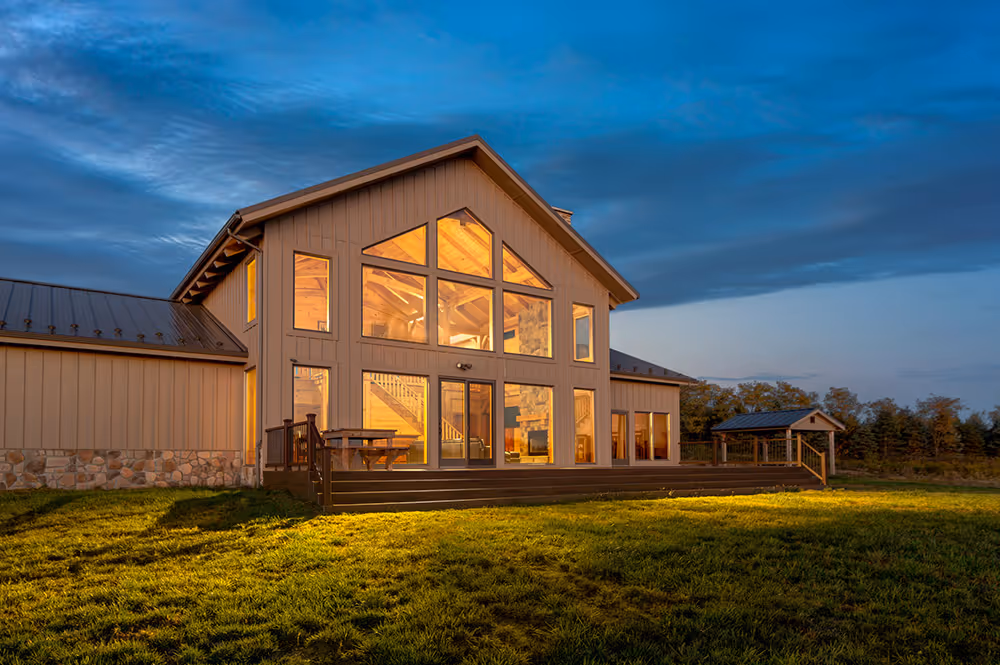 A modern two-story house with large windows reflecting warm light during twilight, surrounded by green grass and trees. The architectural design features a peaked roof and a spacious patio area, enhancing its inviting appearance.