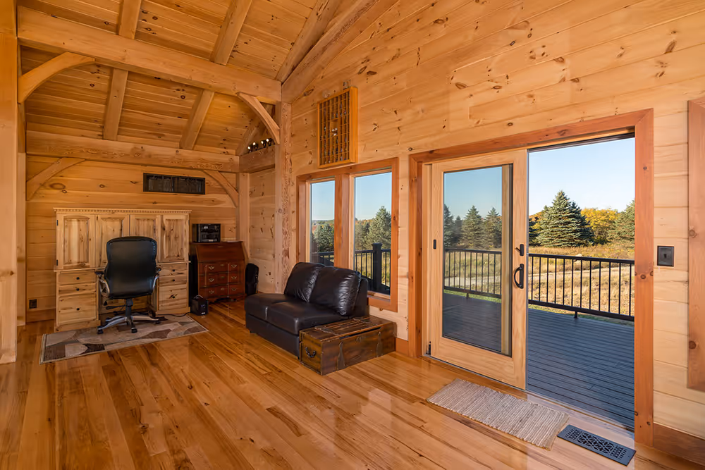 A cozy interior of a wooden cabin featuring a leather sofa, a desk chair, and large glass doors leading to a scenic view outside. The warm wooden walls and flooring add to the inviting atmosphere.