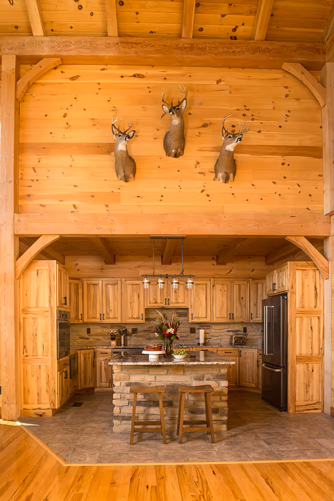 A rustic kitchen featuring wooden cabinetry, a stone countertop island with a vase of flowers, and three mounted deer heads on the wood-paneled wall above. Natural light enhances the warm tones of the wood and flooring.