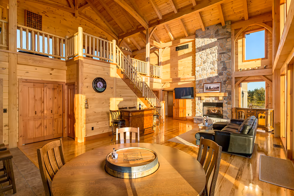 A spacious living area in a wooden cabin featuring high ceilings, large windows letting in sunlight, a stone fireplace, and a wooden dining table at the center with chairs. A staircase leads to the upper level.