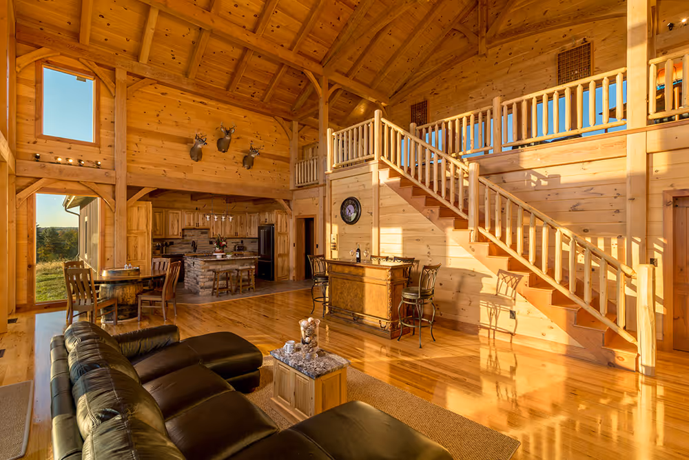 A spacious log cabin interior featuring wooden walls and ceilings, with large windows allowing natural light. A comfortable black sofa faces a wooden bar, and a staircase leads to a loft area.