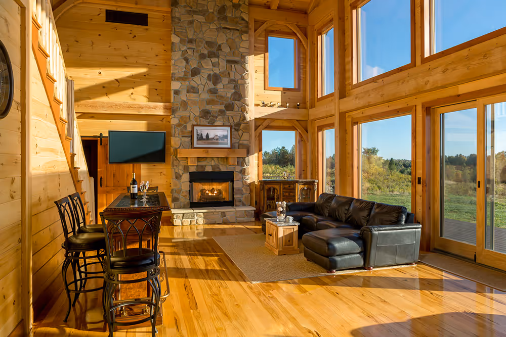 A cozy living room featuring a stone fireplace, large windows letting in natural light, a black leather sofa, and wooden flooring. A small table with two chairs is positioned near the fireplace, and a television is mounted on the wall.