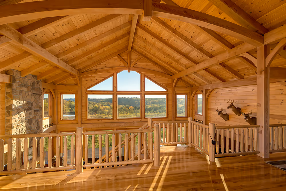 An expansive interior of a rustic wooden cabin featuring vaulted ceilings and tall windows, showcasing scenic views of rolling hills. The wooden balustrade and natural sunlight create a warm, inviting atmosphere.