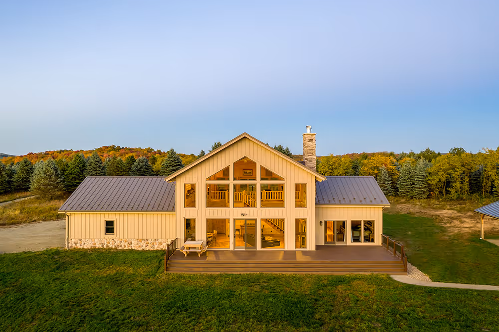 A modern two-story house with a light wood exterior and a metal roof, surrounded by lush greenery and trees. Large glass windows showcase the interior, while a wooden deck invites outdoor relaxation. The scene is serene with a clear sky above.
