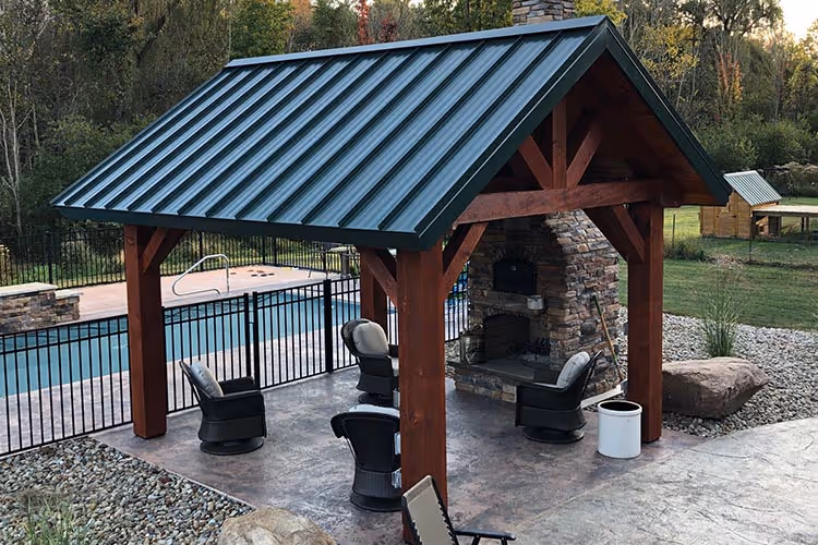 A rustic pavilion with a metal roof and wooden supports, featuring comfortable seats underneath. A swimming pool is visible beyond a black fence, surrounded by lush greenery and stone paths.