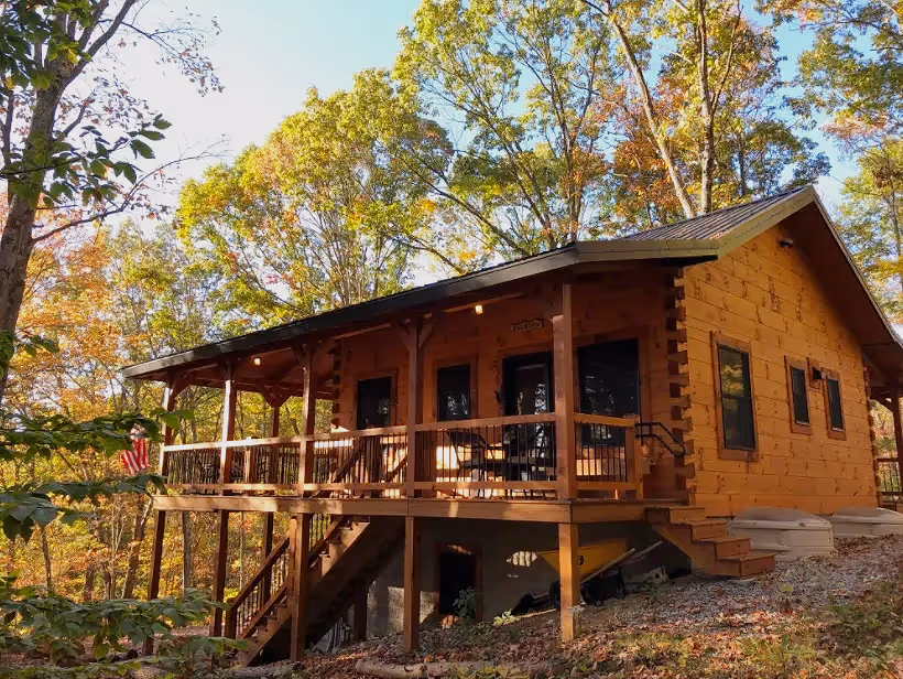 Rustic rental log cabin in a wooded setting featuring a covered elevated porch, outdoor seating, and stairs surrounded by autumn foliage.