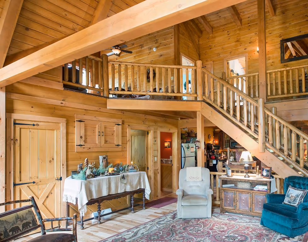 A cozy wooden cabin interior featuring a large open space with a high ceiling. Stairs lead to an upper level with a wooden railing. A table is adorned with decor, and comfortable seating options are arranged on a patterned rug.
