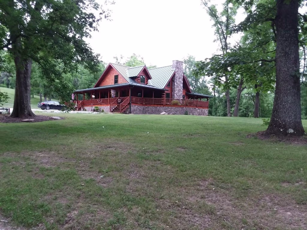 A large rustic log cabin with a green metal roof surrounded by trees, set in a grassy area. The cabin features a spacious porch and a stone chimney, providing a cozy natural retreat.
