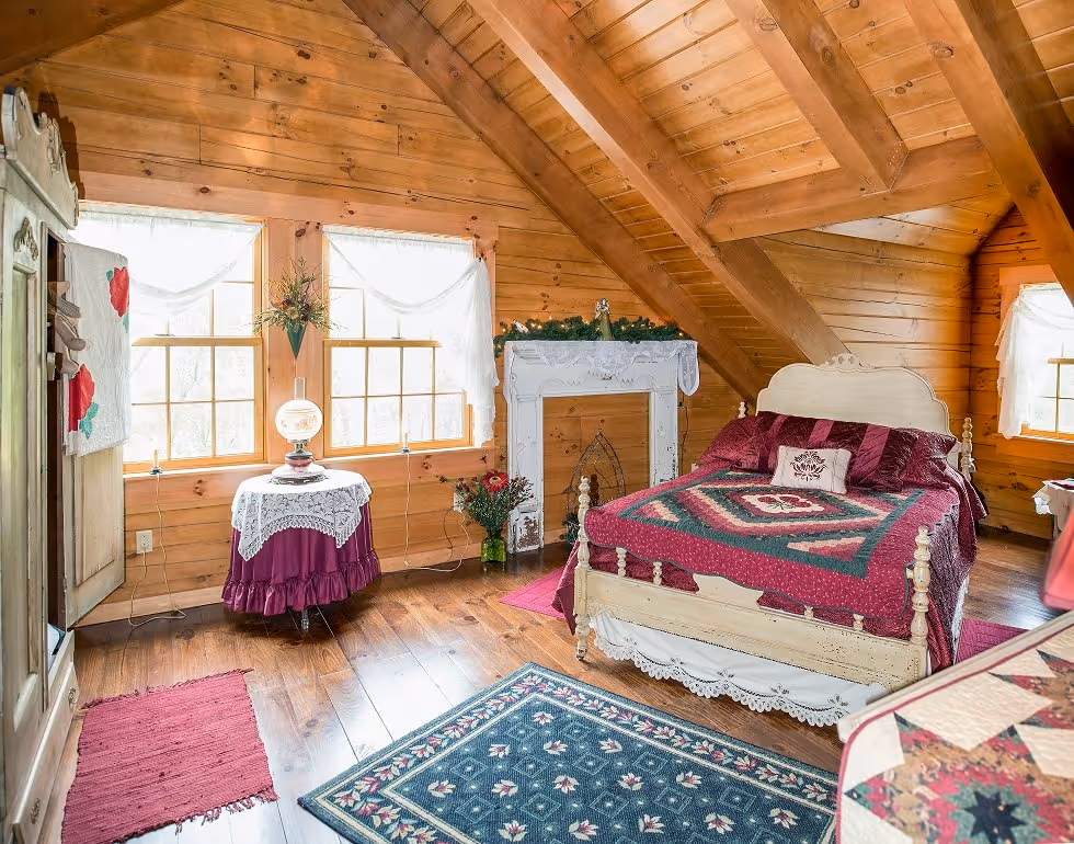 A cozy attic bedroom featuring wooden walls and ceiling beams. A white bed with a burgundy quilt is positioned beside a decorative fireplace. Near the window, a round table with a lamp and a potted plant complements the inviting atmosphere.