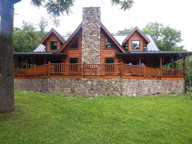 A large rustic log cabin with a stone chimney and a wraparound wooden porch, surrounded by lush green grass and trees. The cabin features multiple peaked roofs and several large windows, creating a cozy and inviting appearance.
