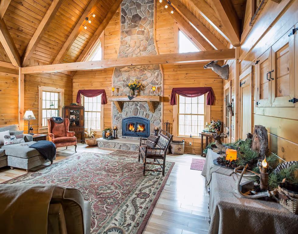 A cozy living room with wooden beams and stone fireplace, featuring a plush gray sofa, a burgundy armchair, and a decorative rug. Windows allow sunlight to fill the space, enhancing the warm cabin atmosphere.