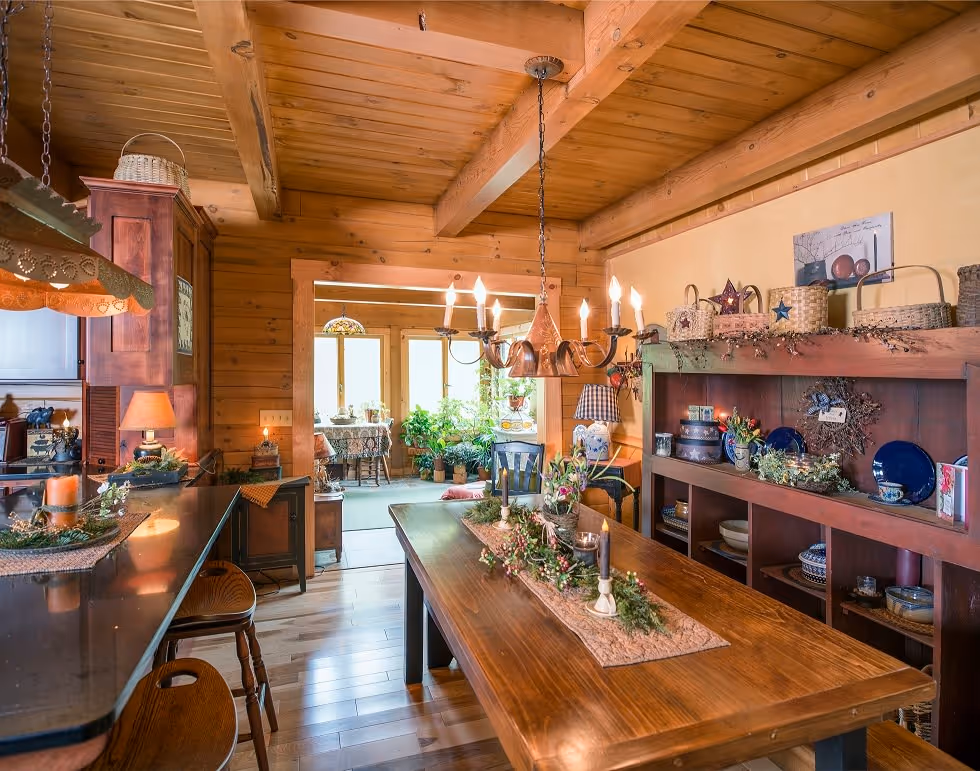 A warm, rustic kitchen featuring wooden beams and furniture. A long dining table with a decorative centerpiece sits in the foreground. The space is filled with potted plants and shelves displaying dishes, creating a cozy atmosphere.