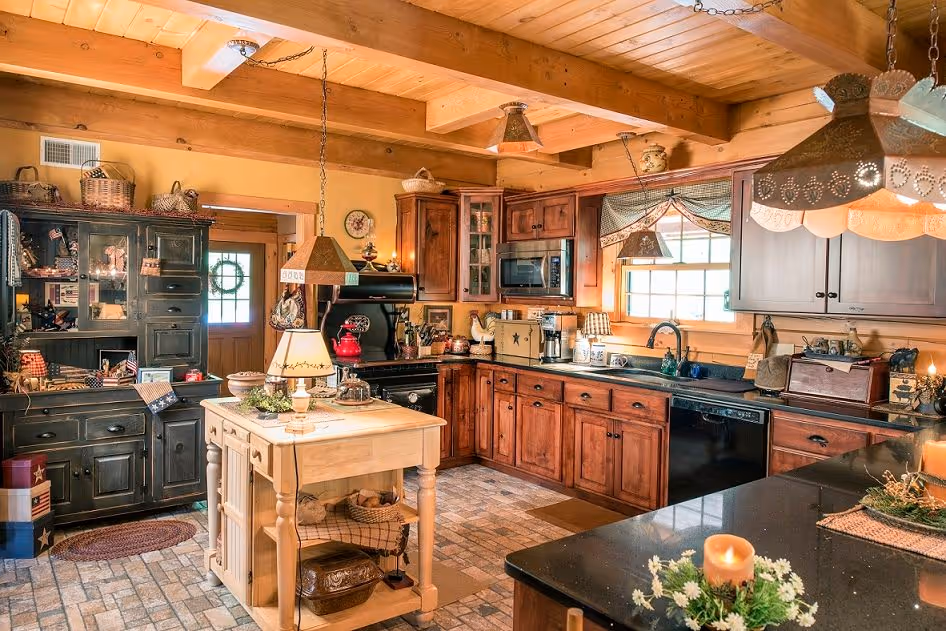 A rustic kitchen featuring wooden cabinets and a black countertop, with a central island adorned with decorative items. Natural light streams through a window, illuminating a cozy ambiance complemented by various kitchen appliances and decor.