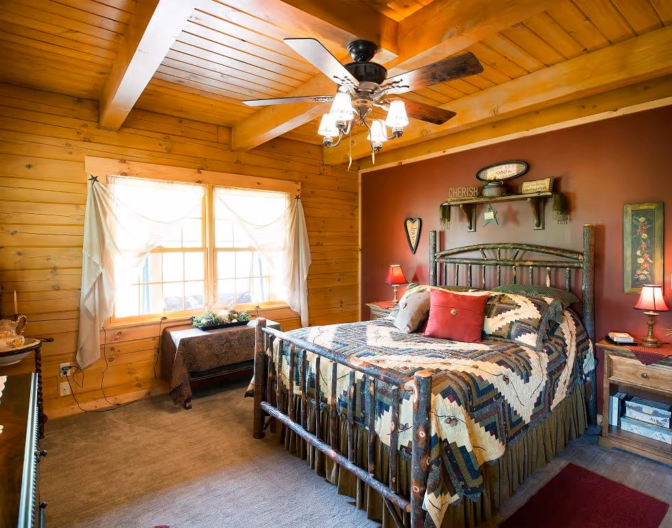 A cozy bedroom featuring a wooden bed frame with a patchwork quilt, two bedside tables with lamps, and a rustic decor. Large windows let in natural light, and walls are made of warm-toned wood. The atmosphere is inviting and tranquil.